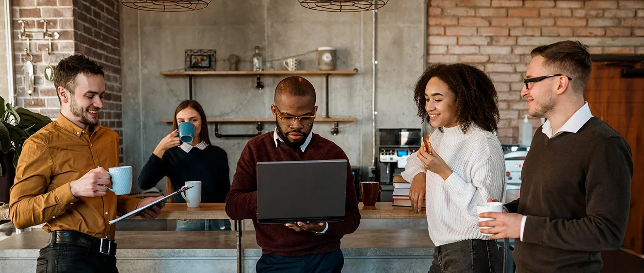Group of people standing up with mugs looking at man with laptop