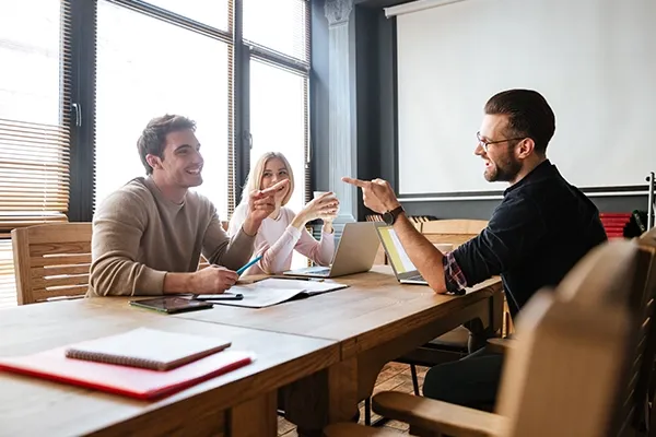 Three people in a meeting room pointing at each other